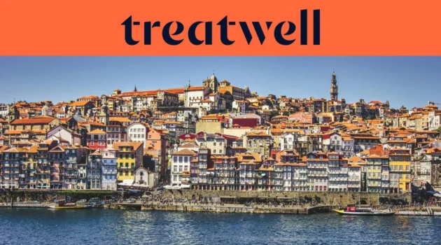 El paseo fluvial de la Ribeira en Oporto, con hileras de casas de colores y tejados de teja que ascienden por la ladera desde el río Duero, y la torre de los Clérigos en el horizonte bajo un cielo azul despejado.