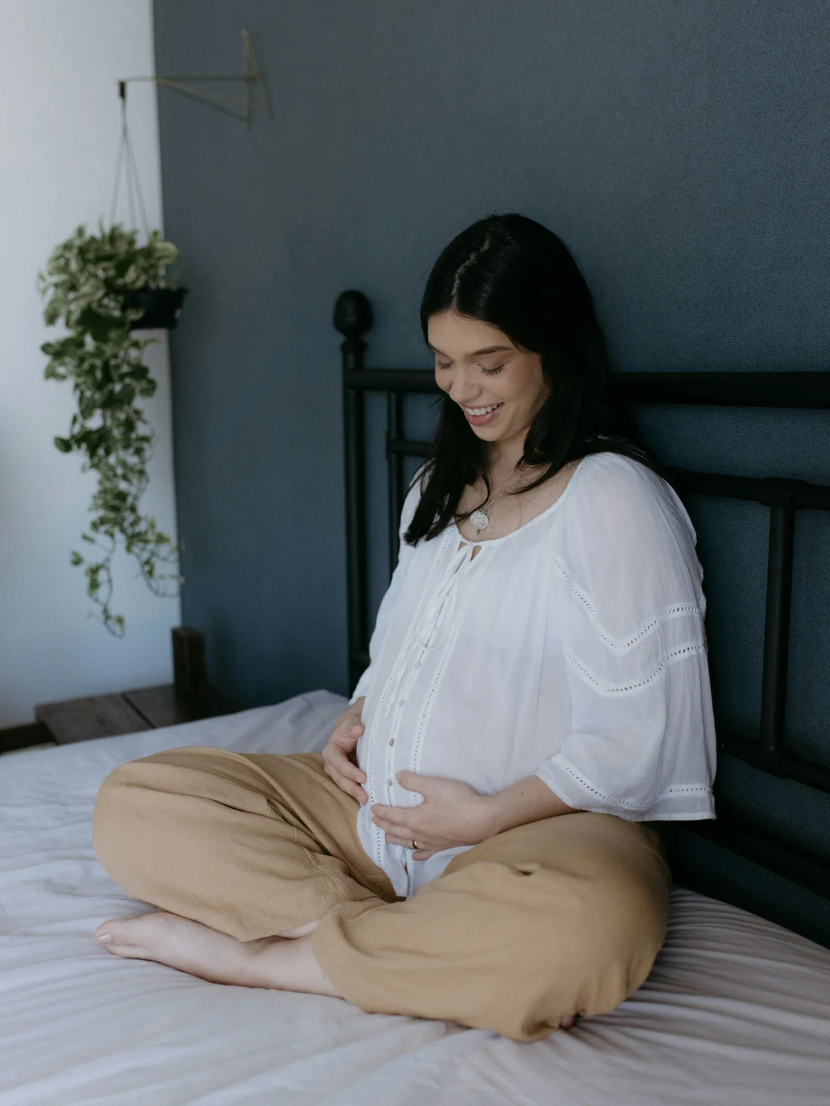 Una mujer embarazada sentada en una cama con las piernas cruzadas, con camisa blanca y pantalón beige, las manos sobre la barriga y sonriendo hacia abajo