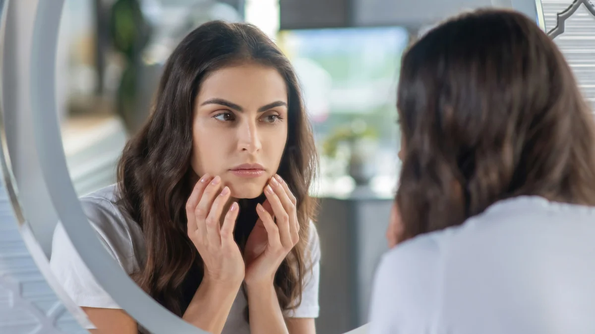 Una mujer de pelo largo y oscuro con las cejas cuidadas y bien definidas se mira al espejo con las manos apoyadas suavemente en el rostro