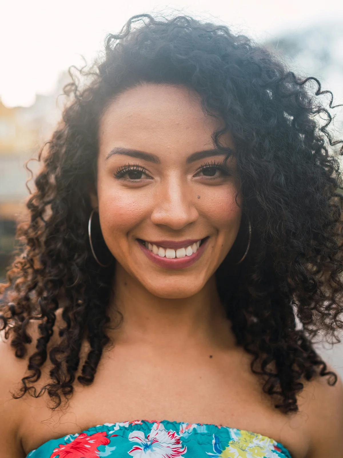 Una mujer con las cejas perfectamente definidas sonríe a la cámara, con un top de flores tipo bandeau, al aire libre con la luz del atardecer
