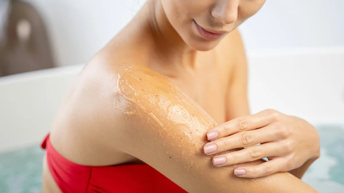 Primer plano de una mujer en bikini rojo aplicándose un exfoliante en la parte alta del brazo en un baño luminoso y ordenado
