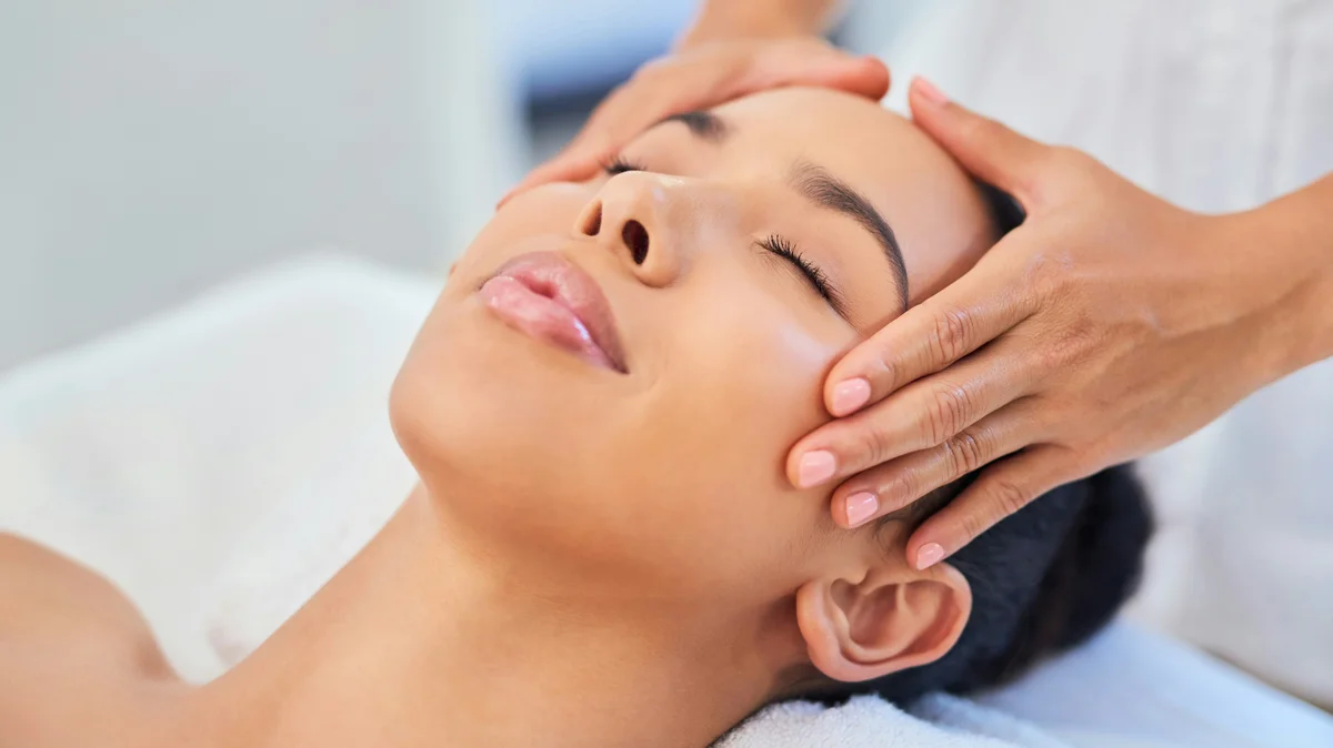 Una mujer tumbada en un centro de estética mientras una esteticista le apoya suavemente las manos en la cabeza durante un tratamiento facial