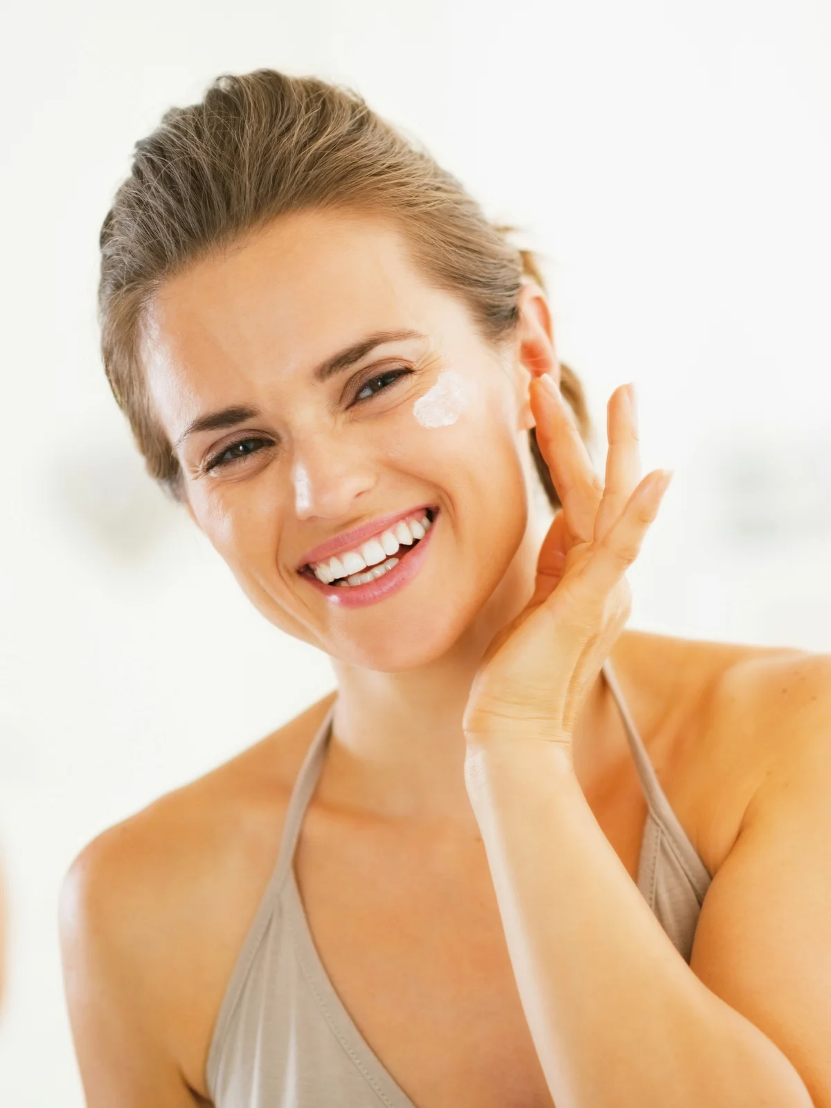 Primer plano de una mujer sonriente con el pelo recogido que se aplica protección solar en el rostro
