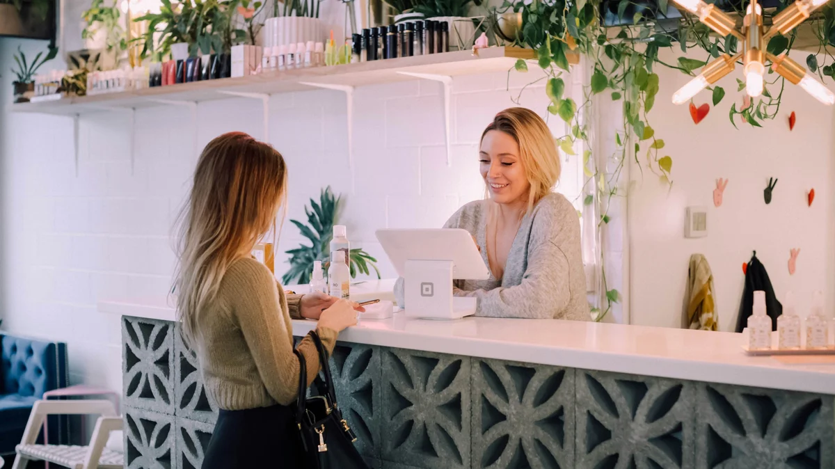 Una mujer está en el mostrador de un centro mientras una recepcionista le reserva su próximo tratamiento; el centro está ordenado, con un mostrador blanco, estanterías de productos bien colocadas y plantas y flores como decoración