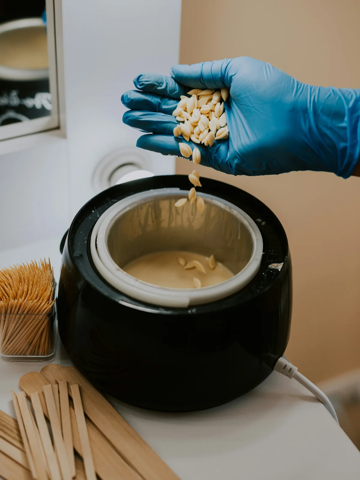 Primer plano de la mano enguantada de una esteticista mezclando perlas de cera caliente en un bote, lista para la cita en cabina