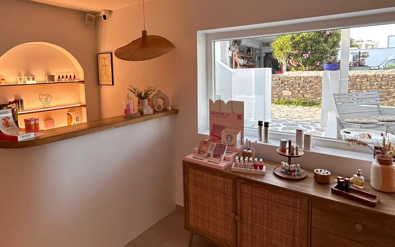 El interior de BeautiQ Esthetics and More en Paros, con una hornacina abovedada suavemente iluminada llena de esmaltes, una lámpara colgante de ratán trenzado y un gran ventanal que da a un muro de piedra y a un jardín soleado.