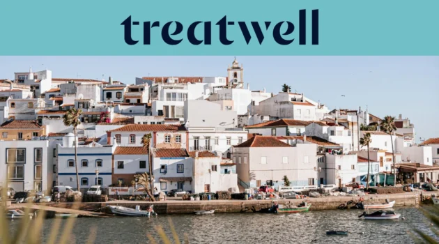 Un pueblo pesquero del Algarve encalado en blanco, con casas de tejados de terracota que ascienden por una ladera sobre una ría en calma, pequeñas barcas de pesca amarradas junto al muelle, palmeras y el campanario de una iglesia bajo un cielo azul despejado.