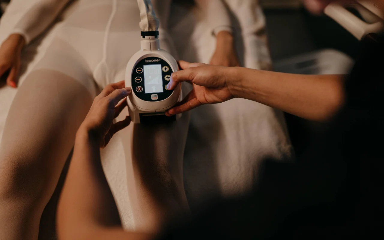 Un tratamiento de remodelación corporal en Beauty Room Romina Bongiovanni, con una profesional que aplica un dispositivo icoone sobre la pierna de una clienta con mono blanco de tratamiento, en una luz cálida y tenue.