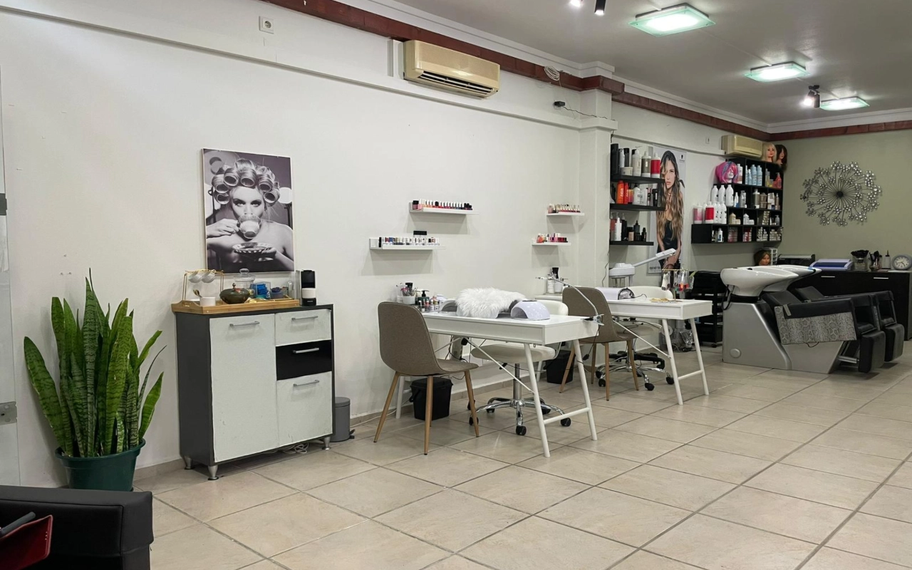 Una peluquería y salón de belleza con dos mesas de manicura blancas y sillas color topo, un lavacabezas y estanterías de productos a lo largo de la pared, una alta sansevieria en un rincón y una gran fotografía en blanco y negro de una mujer con rulos.