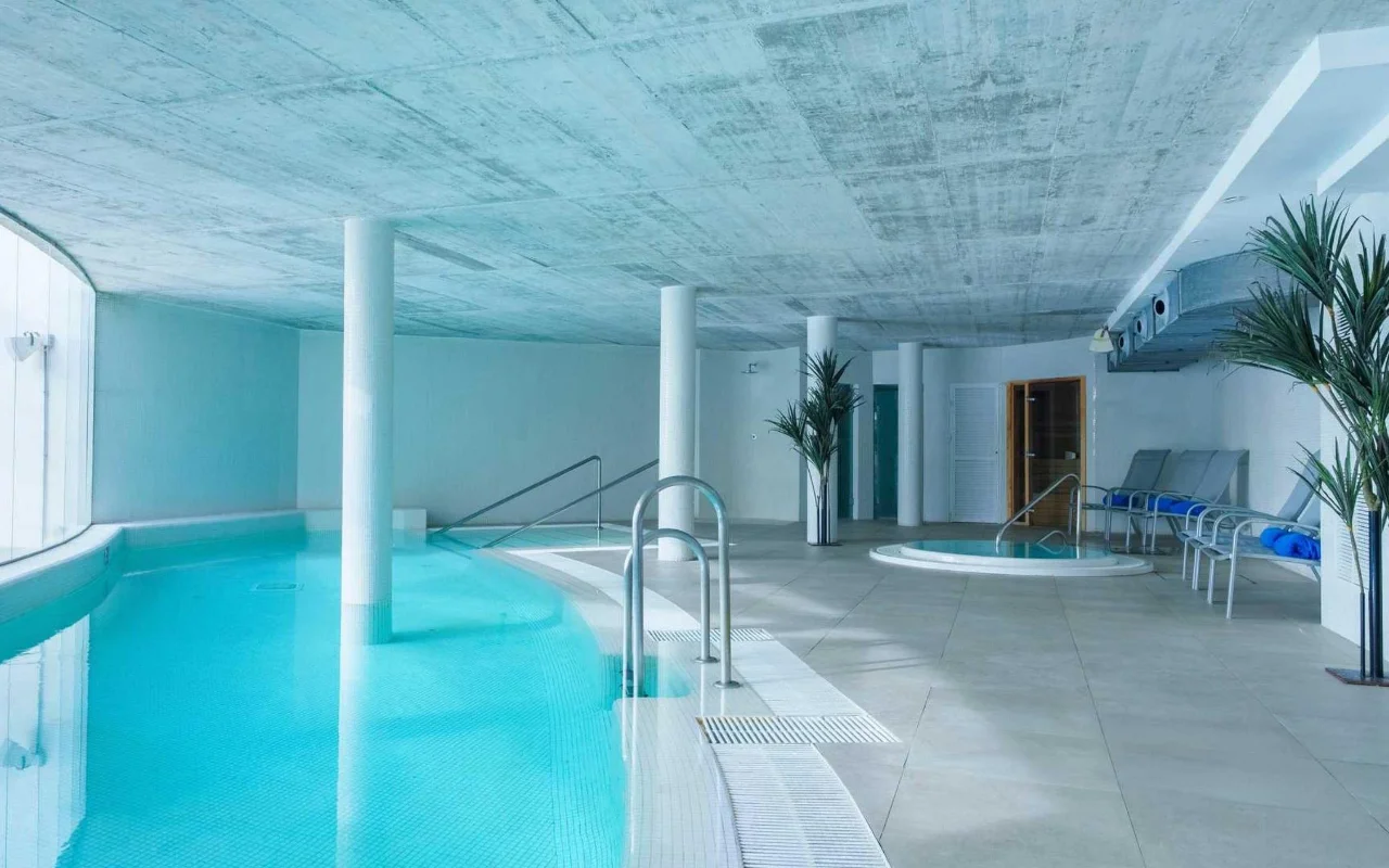 La piscina interior de spa en Benalmádena Beach, con una piscina turquesa curva, un techo de hormigón visto, un pequeño jacuzzi y tumbonas entre palmeras altas en macetas.
