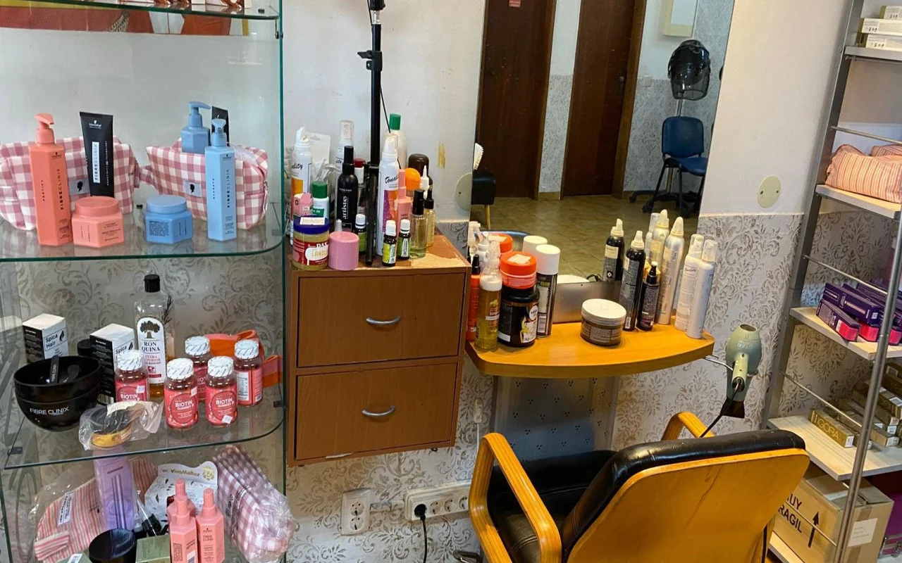 El puesto de un peluquero con un espejo de pared y una encimera de madera repleta de productos de peinado, una vitrina de productos de cuidado capilar y complementos al lado, y un clásico sillón de peluquería en madera y negro.
