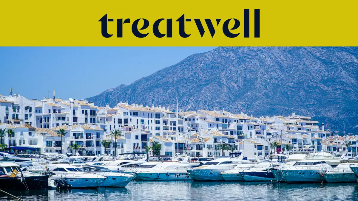 El puerto deportivo de Puerto Banús, en Marbella, rodeado de edificios blancos y yates amarrados con las montañas al fondo, bajo una banderola amarilla de Treatwell.