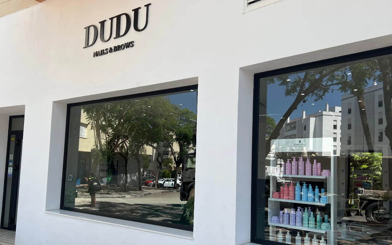 La fachada blanca de Dudu Nails and Brows en Estepona, con su nombre en letras oscuras sobre amplios escaparates que muestran filas de productos para el cabello.