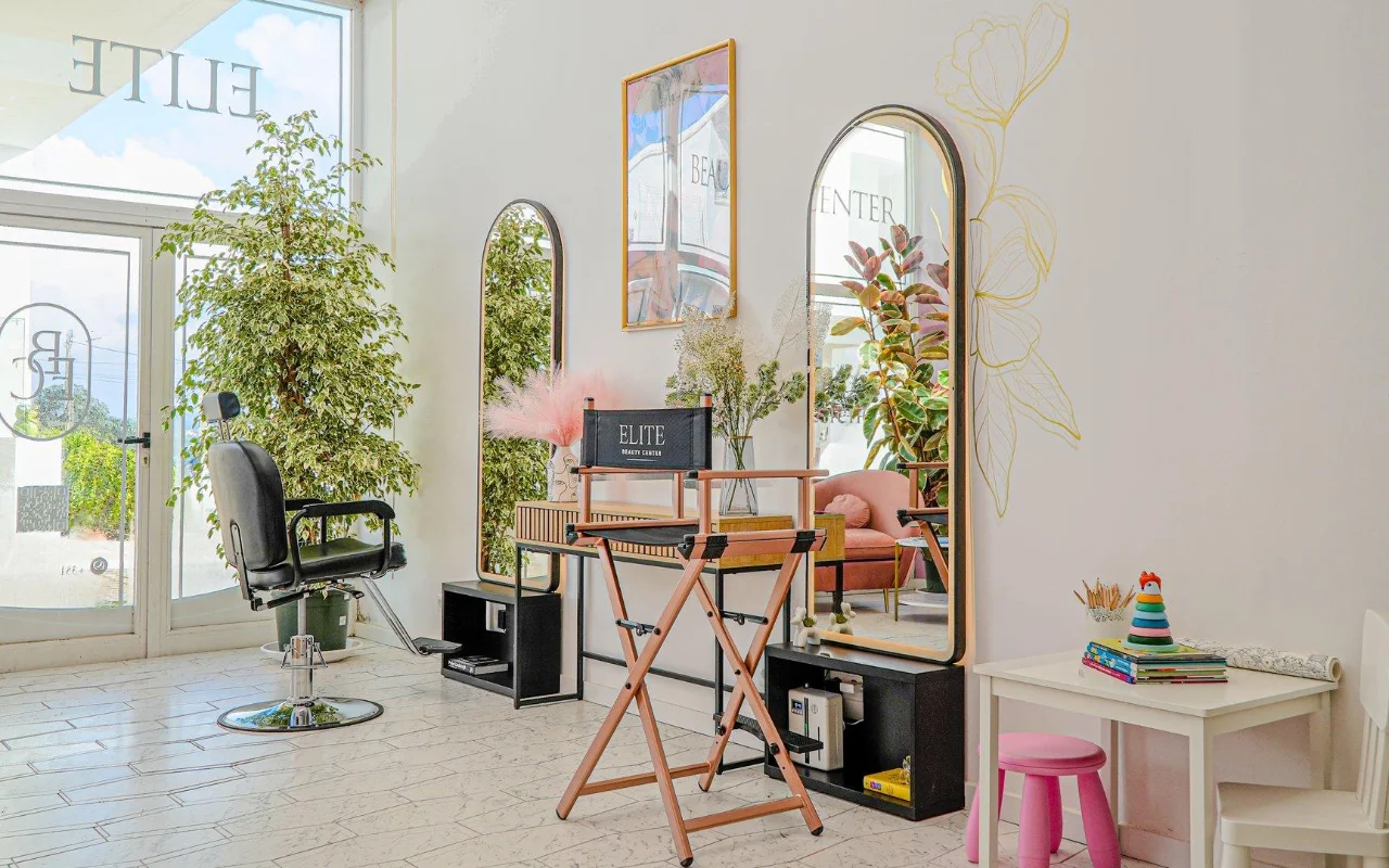 Un salón de belleza luminoso y despejado, todo en blanco, con espejos arqueados de marcos dorados, un sillón de peluquería negro y una silla plegable de director, altas plantas verdes, toques de rosa y un pequeño rincón infantil junto a la ventana.