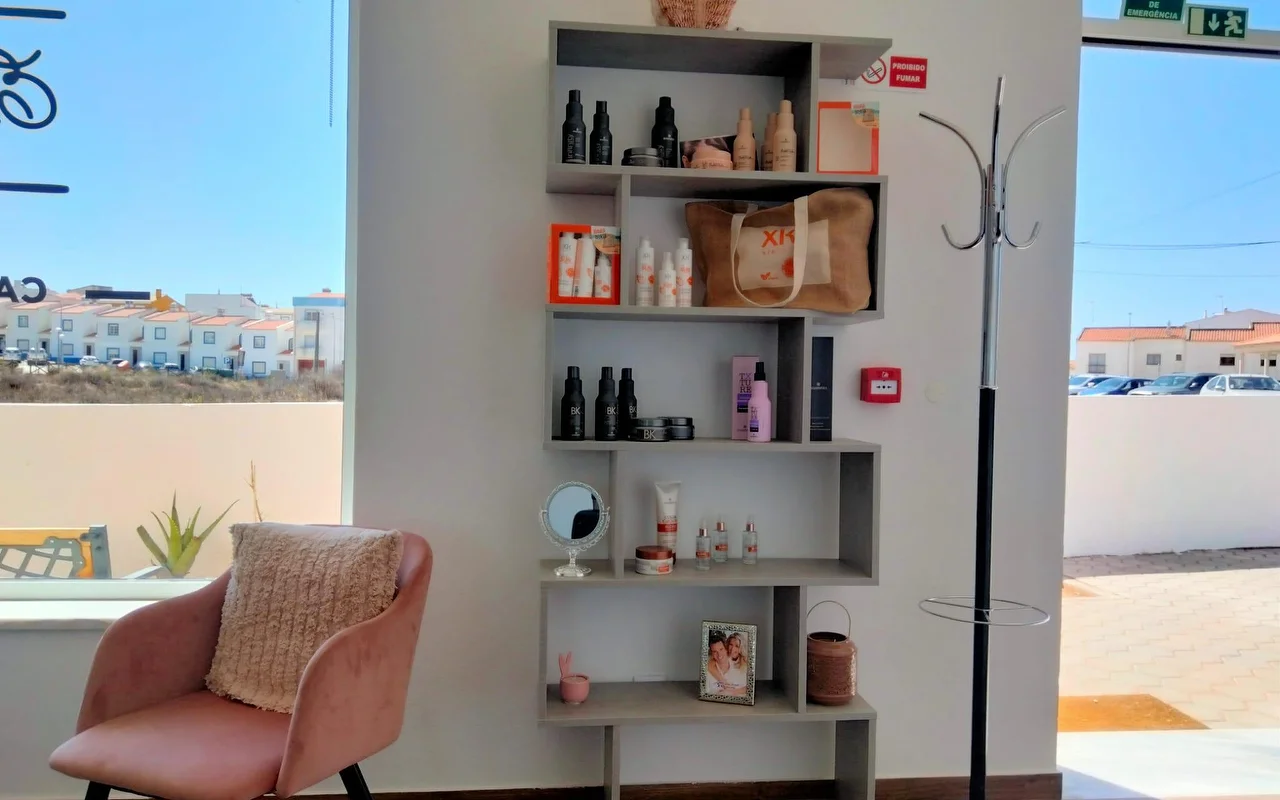 Un rincón de salón junto a un gran ventanal, con una estantería gris escalonada llena de productos para el pelo y una foto enmarcada, un sillón de terciopelo rosa empolvado con un cojín, y vistas a casas bajas encaladas bajo un cielo azul.