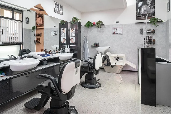 Sillones de barbero vintage y lavacabezas en Fabio Desole Acconciature Maschili en Sassari, con retratos enmarcados en las paredes.