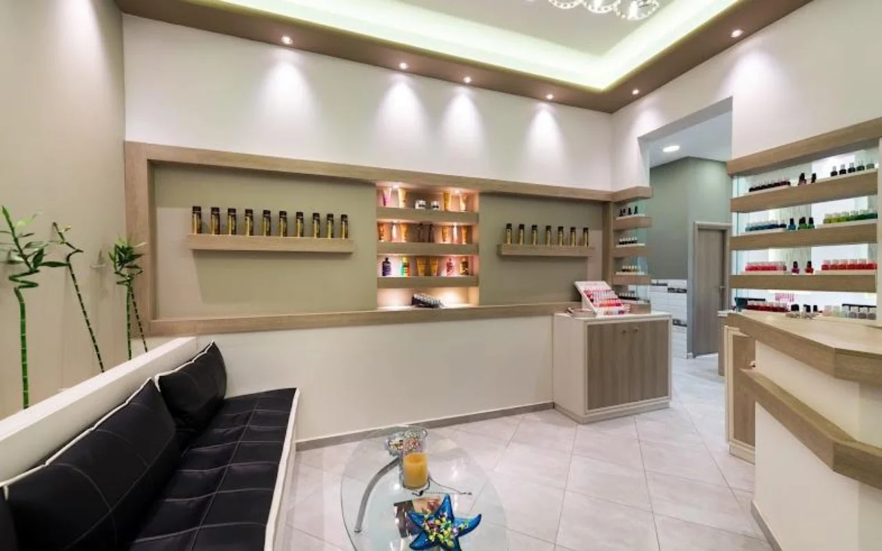 La recepción en tonos crema de Fabulous Nails and More en Fira, Santorini, con un banco de piel negra, una mesa de cristal, estanterías retroiluminadas con productos de belleza y una luz de techo suave.