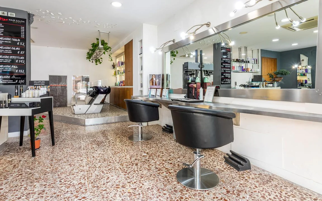 El interior del Hair Salon Luigi Zito, con suelo de terrazo, dos sillones de peinado negros frente a un largo espejo, listas de precios en pizarra en la pared, un expositor de productos y un lavacabezas al fondo.