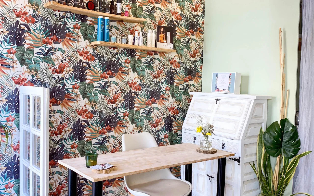 Un puesto de peinado en Shangara Lora Organics en Marbella, con un llamativo papel pintado tropical, una mesa de madera, un antiguo escritorio blanco y productos capilares orgánicos en estantes flotantes.