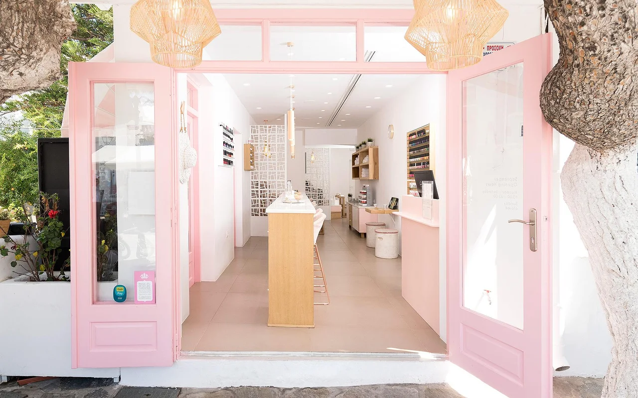 La entrada enmarcada en rosa de Vivian Nail Boutique en Naoussa, Paros, con las puertas abiertas, lámparas colgantes trenzadas y un mostrador de madera, en un edificio cicládico encalado junto a un árbol.