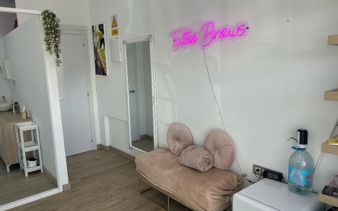 Un espacio de tratamiento en YG Beauty Estudio en Torremolinos, con un letrero de neón rosa que dice «Tattoo Brows», un espejo de cuerpo entero y un diván beige acolchado.