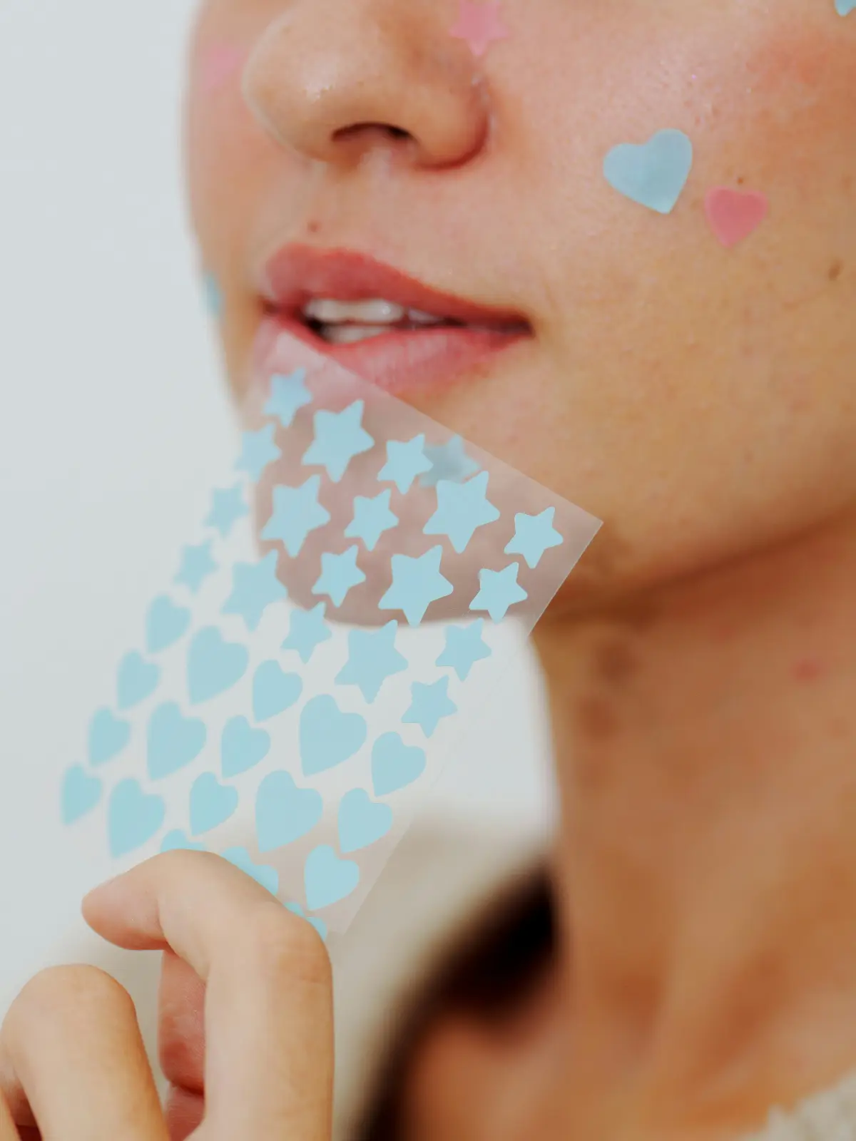 Primer plano del rostro de una mujer con varios parches hidrocoloides con forma de corazón y de estrella sobre sus granos