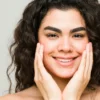 Una joven con la piel limpia y el pelo oscuro y ondulado sonríe a la cámara