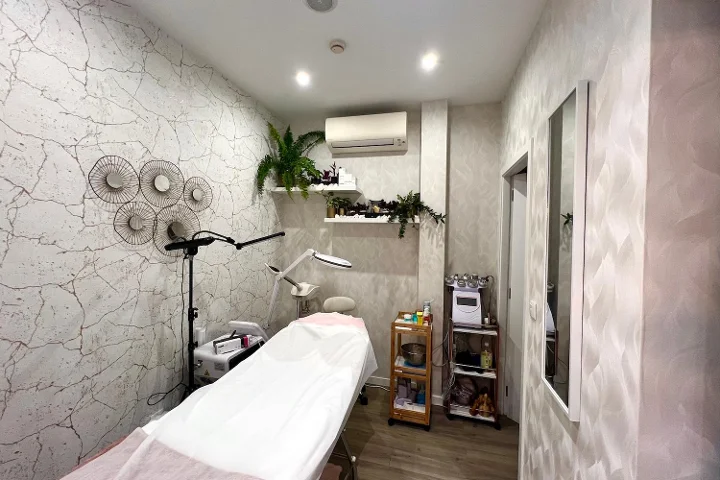 Cabina de estética tranquila con una camilla cubierta, una lámpara de aumento y un vaporizador, un carrito con productos, paredes texturizadas en gris pálido decoradas con discos metálicos y plantas que caen sobre las estanterías.