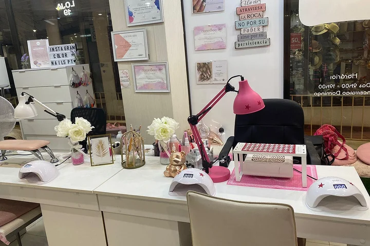 Puesto de manicura de un salón de uñas con mesas blancas, una lámpara rosa fuerte y lámparas LED, rosas blancas y certificados enmarcados en la pared, con el nombre Color Nails PT visible en el escaparate junto al horario.