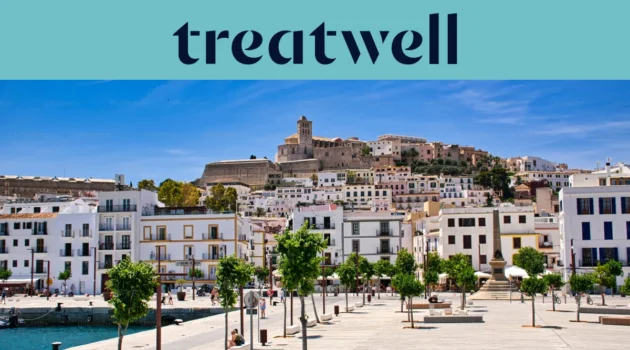 Vista soleada sobre Ibiza ciudad, con edificios encalados en blanco que suben hasta la catedral y las antiguas murallas de Dalt Vila en la colina. Un paseo marítimo arbolado atraviesa el primer plano bajo un cielo azul limpio.