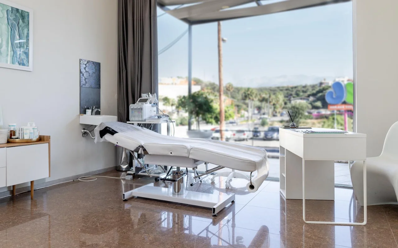 Una sala de tratamiento blanca e impoluta con una camilla de belleza reclinable, equipos de skincare sobre un carro, obras enmarcadas y una gran ventana con vistas a árboles, una carretera y colinas a lo lejos.