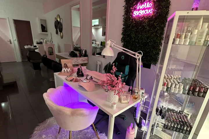 Puesto de belleza iluminado en rosa, con un cartel de neón 'hello gorgeous' sobre una pared vegetal, un sillón crema con forma de concha, una mesa blanca con brochas y flores de cerezo, y una vitrina llena de esmaltes de colores.
