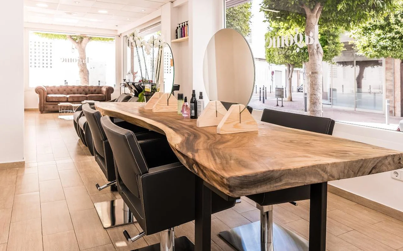 Un salón de peluquería luminoso construido en torno a un largo mostrador de madera de canto natural, con sillas de peluquería negras. Un espejo redondo y expositores de madera para productos están sobre el mostrador, un sofá Chesterfield de cuero marrón espera al fondo, y ventanas altas dan a una calle arbolada.
