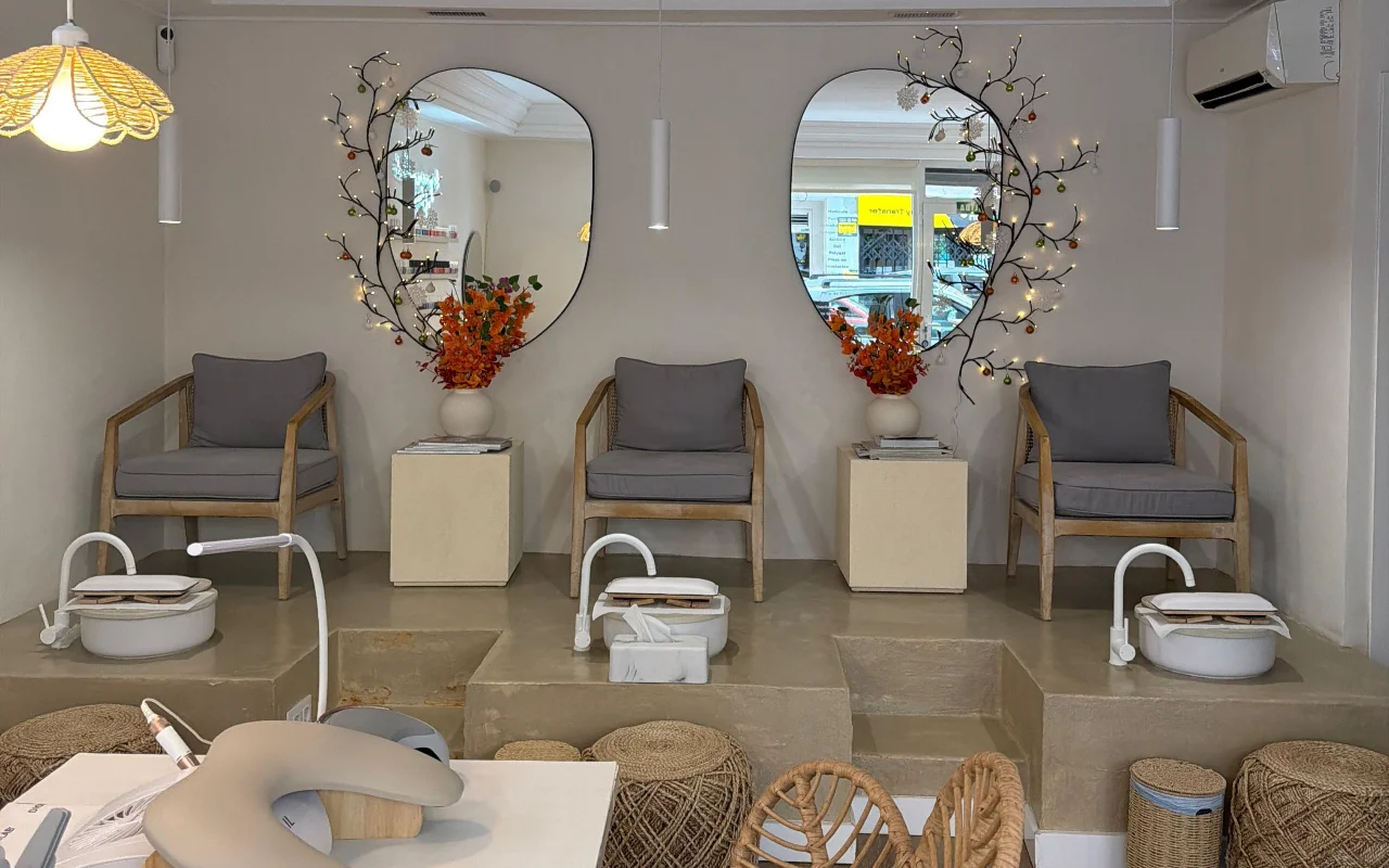 Un salón de uñas moderno y luminoso con dos sillones de pedicura sobre una plataforma de cemento elevada, cada uno con una bañera blanca para los pies y un sillón gris acolchado. Dos espejos con formas orgánicas, decorados con ramas iluminadas y flores anaranjadas, cuelgan por encima, con una estación de manicura en primer plano.