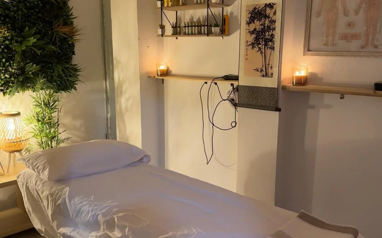 Una sala de tratamientos con luz tenue con una camilla de masaje vestida en lino blanco. Velas encendidas descansan en estantes de pared de madera junto a un pergamino en tinta de bambú y un cuadro enmarcado con un mapa de acupuntura, con una planta frondosa y una lámpara tejida encendida en la esquina.