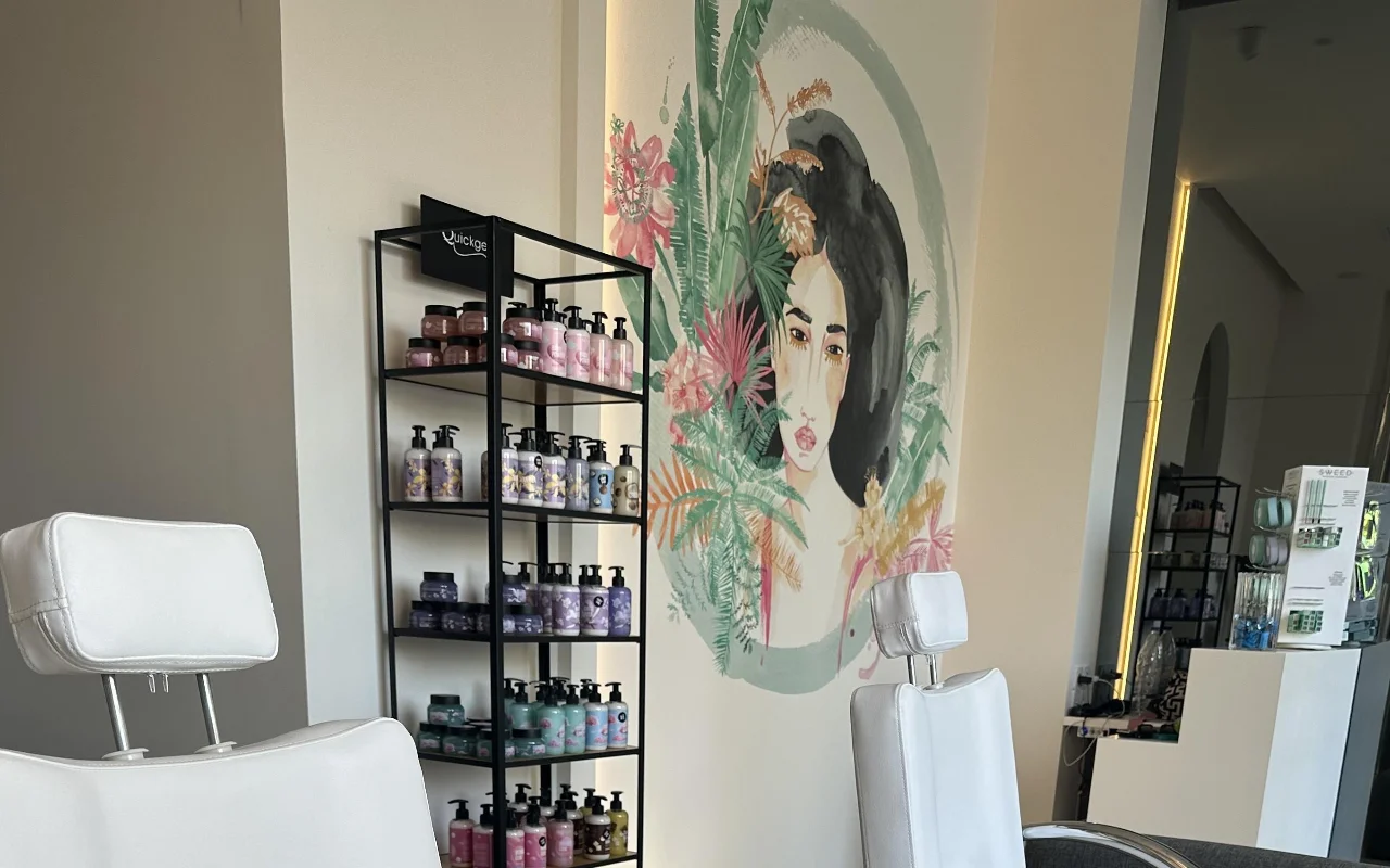 Un rincón de estudio con dos sillones de tratamiento blancos reclinables, una estantería negra de productos, un espejo retroiluminado, y un mural floral con el rostro de una mujer enmarcado por follaje tropical.
