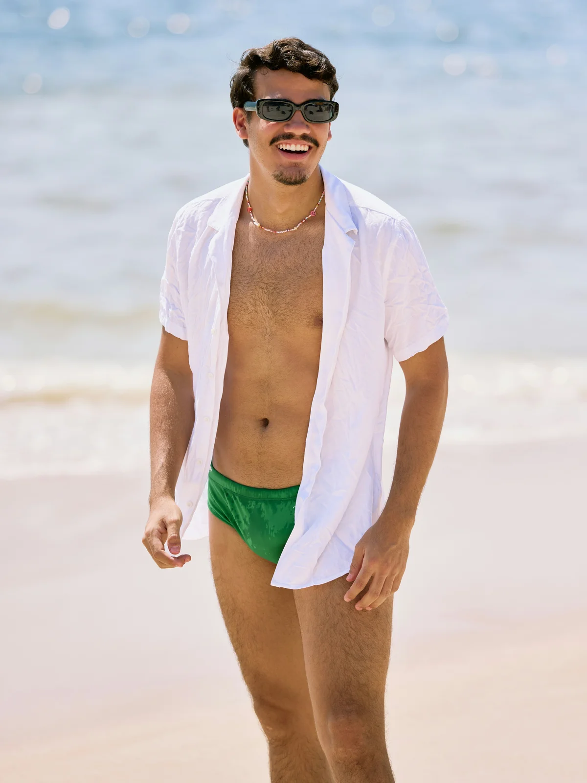 Un hombre sonriente está de pie en una playa en un día soleado; lleva un bañador verde, una camisa blanca abierta y gafas de sol negras