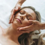 Mujer relajada durante un masaje facial de drenaje linfático suave, con las manos de la profesional deslizándose ligeras por su rostro